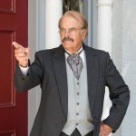 Gene Worthington as Theodore Roosevelt, wearing dark gray suit with light gray vest, gray ascot, and white dress shirt, spectacles, grayish-black mustache and thinning hair, with left hand on hip and right hand, with wedding band on ring finger, pointing away