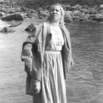 Black and white image of Ilene Evans as Harriet Tubman, a Black woman wearing a light-colored dress and darker jacket with a smock on her head, standing on the edge of a stream with rocks in the background, looking up