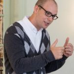 James Froemel as Charles Schulz, with cardigan with light gray and dark gray diamond shapes, white dress shirt, spectacles, crew cut, curled hands with both thumbs pointing up