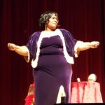 Doris Fields as Bessie Smith, Black woman with black hair, wearing fancy purple outfit fringed in white, both arms stretched out from the elbows, standing in front of a table with a red tablecloth and frame photo, in front of a burgundy stage curtain