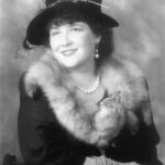Black and white image of Karen Vuranch as Pearl S. Buck, with dark hair and gloved hands folded, wearing a black hat with ornamentation, a dark dress, a light stole, pearl earrings and necklace, and smiling