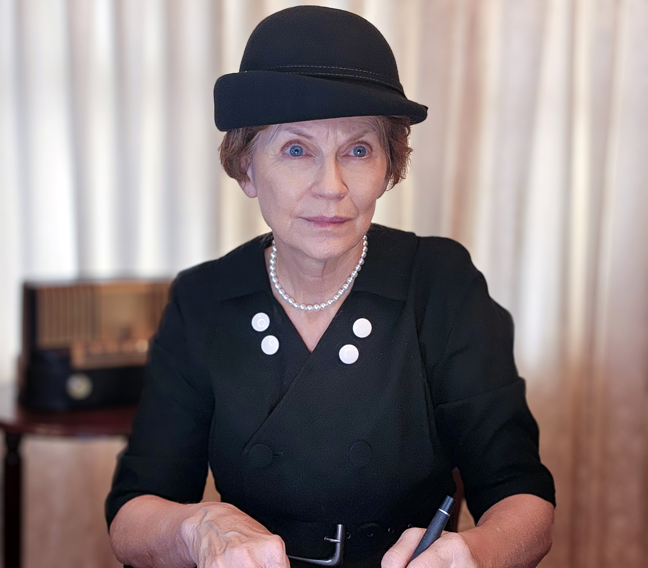 JoAnn Peterson as Frances Perkins, with reddish-brown hair and blue eyes seated with a pen in her hand, wearing a black dress with black and white buttons, a pearl necklace, and a black hat