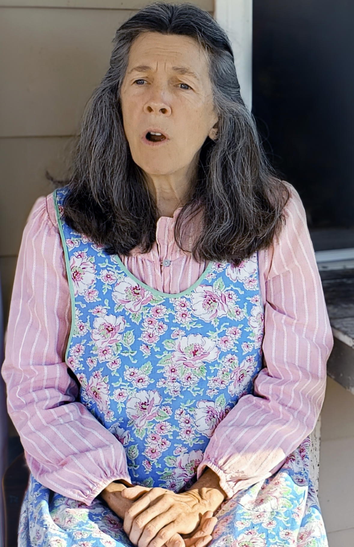 Mary Dailey as a composite character, seated with brown hair and brown eyes singing, wearing a flowery blue smock and pink shirt with white stripes, with hands crossed