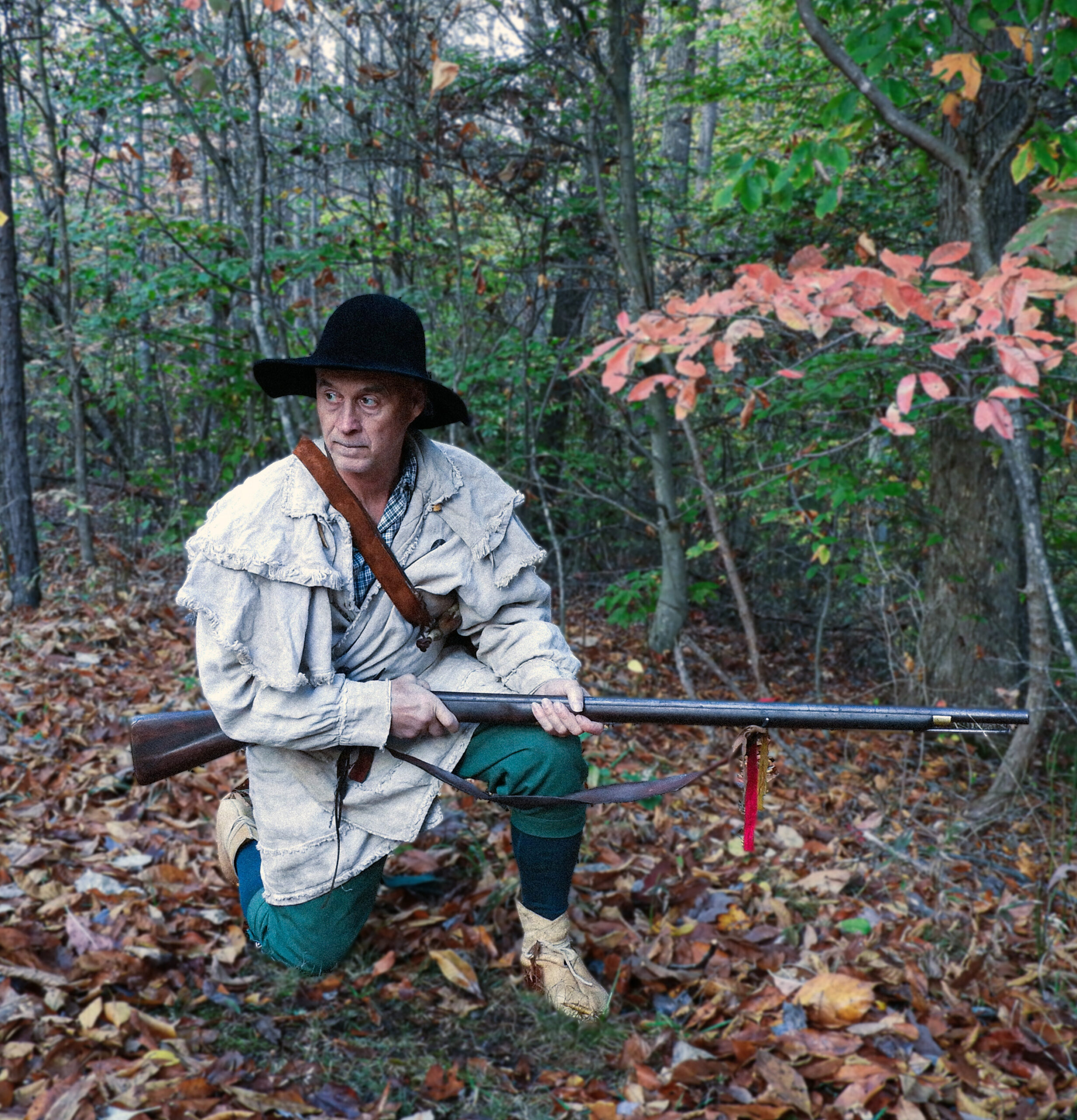 Doug Wood as Thomas Ingles, dressed as a 1700s hunter in the woods with a musket, wearing a black hat, off-white jacket, green britches, and beige shoes