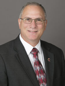 Peter Mansoor Man with thinning gray hair, glasses, dark gray suit coat, abstract red and gray tie, white shirt, and a lapel pin with The Ohio State University logo, smiling at the camera